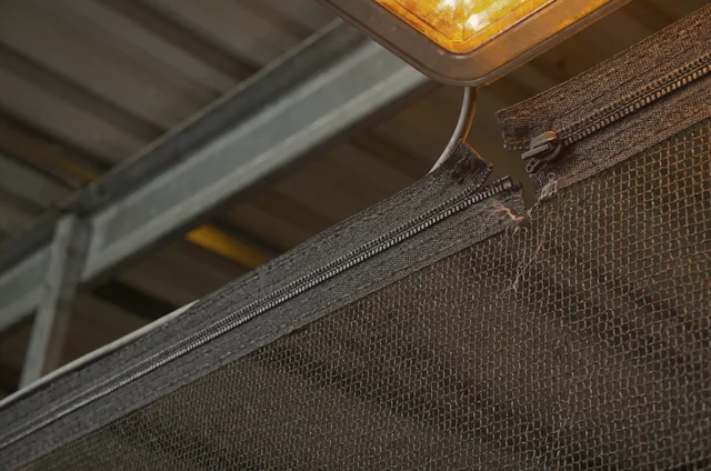 Close-up of a warehouse netting access zip beside a high-bay light, showing separated teeth behind the slider and frayed stitching, an item on a bird netting maintenance checklist for warehouses